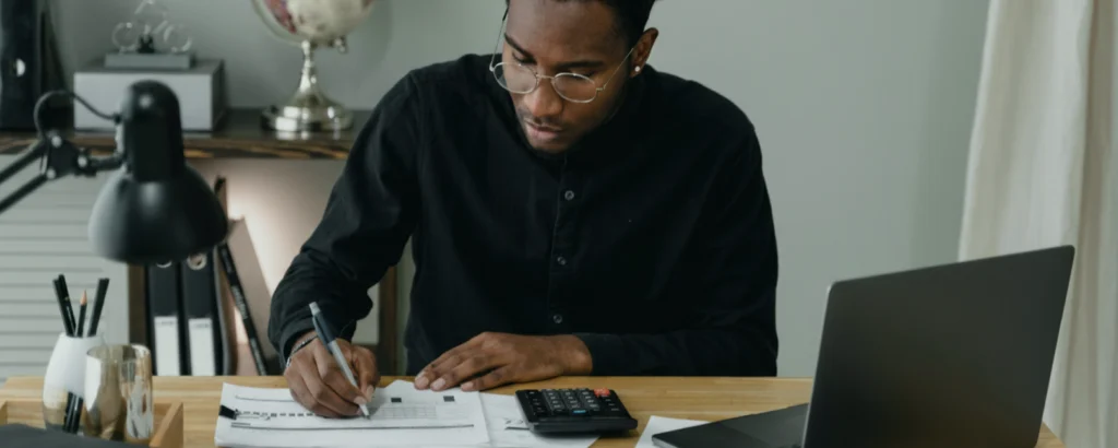 Homme calcule son budget mensuel en utilisant une calculatrice sur smartphone avec fiches de paies et factures.